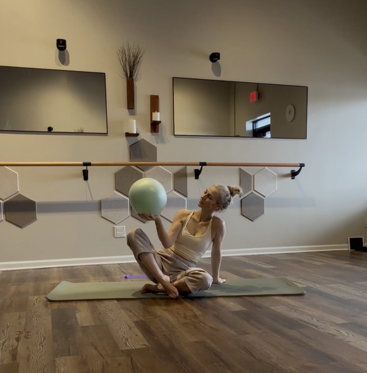 Lift & Stabilize class at Mindful Way Studio Mokena IL — Ania seated on mat holding Pilates ball with barre and hexagon acoustic panels in background